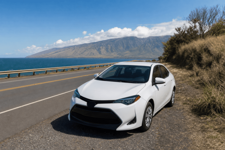 White economy rental car parked on Honoapiilani Highway shoulder in Maui with ocean and mountains behind.